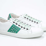 Three quarter front view of low top white and green sneaker. Full grain leather upper with silver studs and white rubber sole.