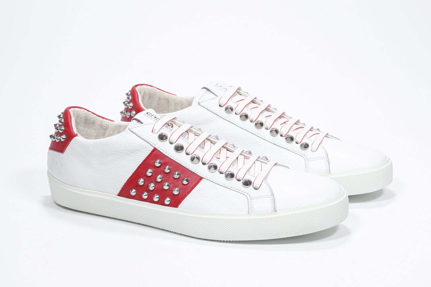 Three quarter front view of low top white and red sneaker. Full grain leather upper with silver studs and white rubber sole.