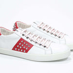 Three quarter front view of low top white and red sneaker. Full grain leather upper with silver studs and white rubber sole.