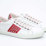 Three quarter front view of low top white and red sneaker. Full grain leather upper with silver studs and white rubber sole.