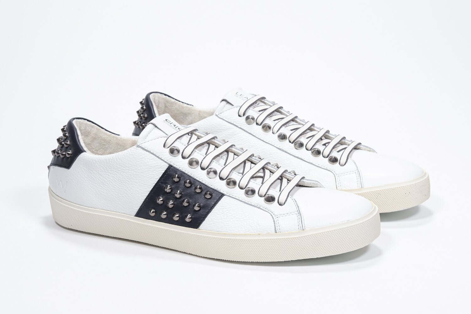 Three quarter front view of low top white and navy sneaker. Full grain leather upper with gunmetal studs and vintage rubber sole.