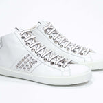 Vista frontale di tre quarti del modello mid top bianco sneaker. Tomaia in pelle con borchie, zip interna e suola in gomma bianca.