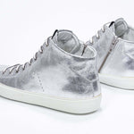 Vue de trois quarts arrière d'un modèle argenté de taille moyenne sneaker avec une tige en cuir avec un logo perforé, une fermeture à glissière interne et une semelle blanche.