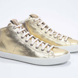 Vue de trois quarts de l'avant de la chaussure de taille moyenne en or sneaker avec une tige en cuir avec un logo perforé, une fermeture éclair interne et une semelle blanche.