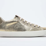 Seitliches Profil des goldenen Low Top sneaker mit perforiertem Kronenlogo auf dem Obermaterial. Obermaterial aus Metallic-Leder und weiße Gummisohle.