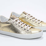 Dreiviertelansicht der Vorderseite des goldenen Low Top sneaker mit perforiertem Kronenlogo auf dem Obermaterial. Obermaterial aus Metallic-Leder und weiße Gummisohle.