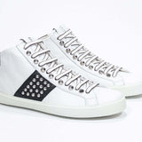 Tre quarti della parte anteriore del modello mid top bianco e nero sneaker. Tomaia in pelle con borchie, zip interna e suola in gomma bianca.