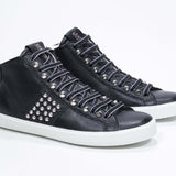 Vista frontale di tre quarti del modello mid top nero sneaker. Tomaia in pelle con borchie, zip interna e suola in gomma bianca.