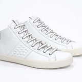 Three quarter front view of mid top white sneaker. Full leather upper with studs, an internal zip and vintage rubber sole.
