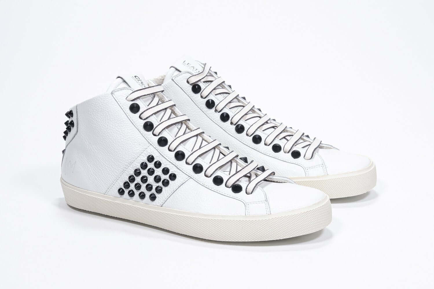 Three quarter front view of mid top white sneaker. Full leather upper with studs, an internal zip and vintage rubber sole.