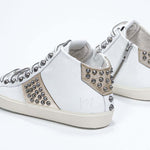 Vue de trois quarts arrière d'un modèle intermédiaire blanc et or métallisé sneaker. Tige en cuir avec clous, fermeture à glissière interne et semelle en caoutchouc vintage.