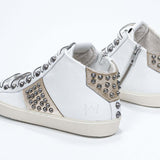 Vue de trois quarts arrière d'un modèle intermédiaire blanc et or métallisé sneaker. Tige en cuir avec clous, fermeture à glissière interne et semelle en caoutchouc vintage.