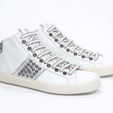 Dreiviertelansicht der Vorderseite von Mid Top weiß und metallisch silber sneaker. Obermaterial aus Vollleder mit Nieten, einem internen Reißverschluss und einer Vintage-Gummisohle.
