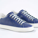Three quarter front view of low top denim blue sneaker with white detailing and perforated crown logo on upper. Full leather upper and white rubber sole.