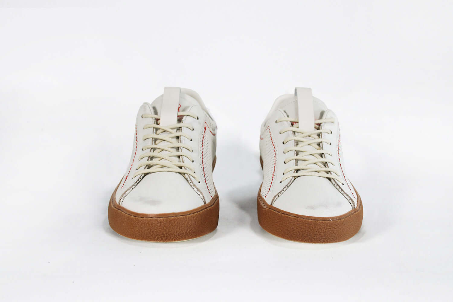 Front view of low top white sneaker. Perforated aged white leather upper with red stitching and dark rubber sole.