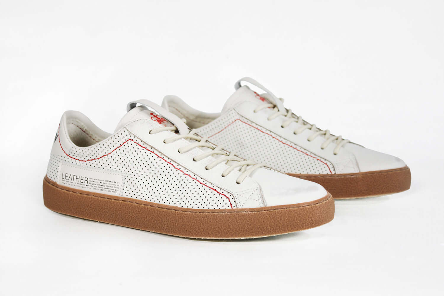 Three quarter front view of low top white sneaker. Perforated aged white leather upper with red stitching and dark rubber sole.
