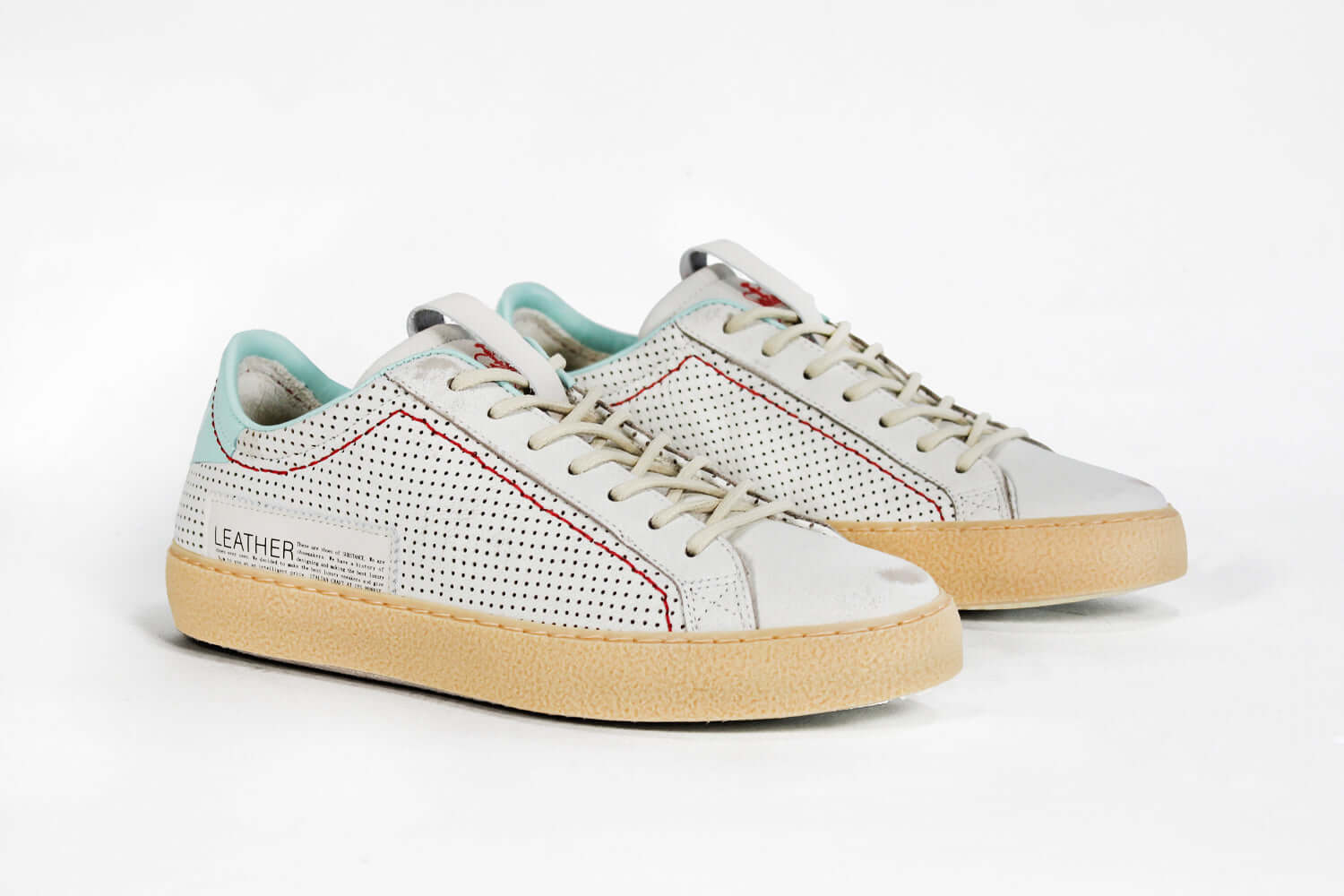 Three quarter front view of low top perforated aged white leather sneaker with pale blue accents, red stitching detail and pale rubber sole.