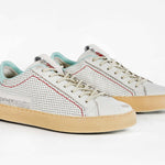 Three quarter front view of low top perforated aged white leather sneaker with pale blue accents, red stitching detail and pale rubber sole.