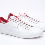 Three quarter front view of low top white sneaker with red detailing and perforated crown logo on upper. Full leather upper and white rubber sole.