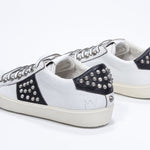 Three quarter back view of low top white and black sneaker. Full leather upper with studs and vintage rubber sole.