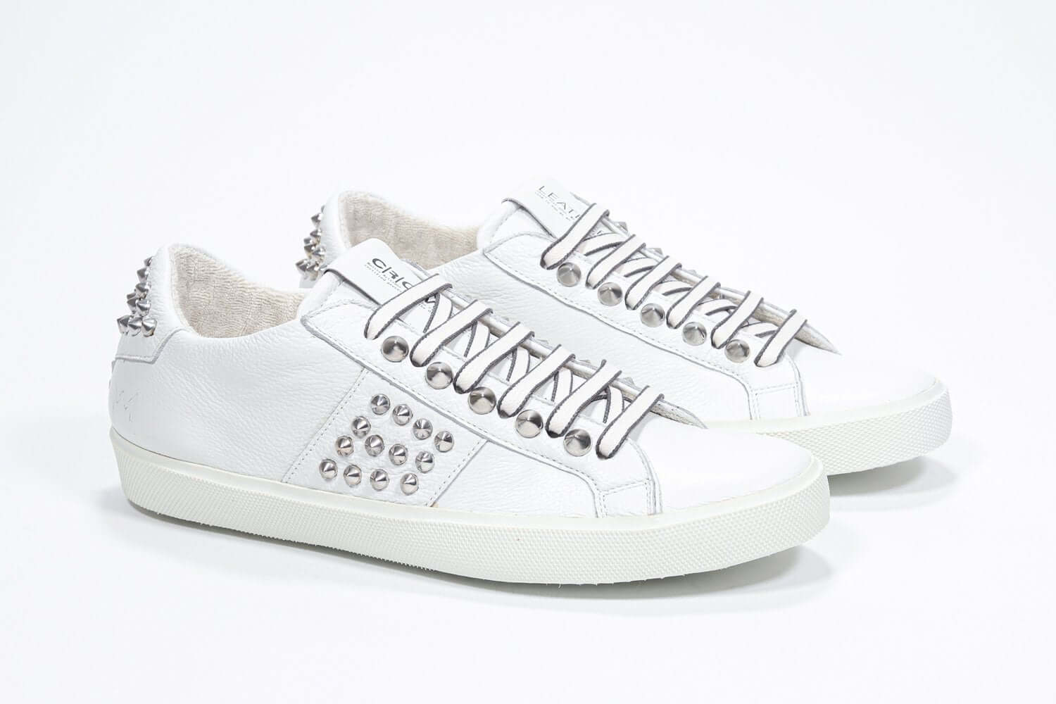 Three quarter front view of low top white sneaker. Full grain leather upper with silver studs and white rubber sole.