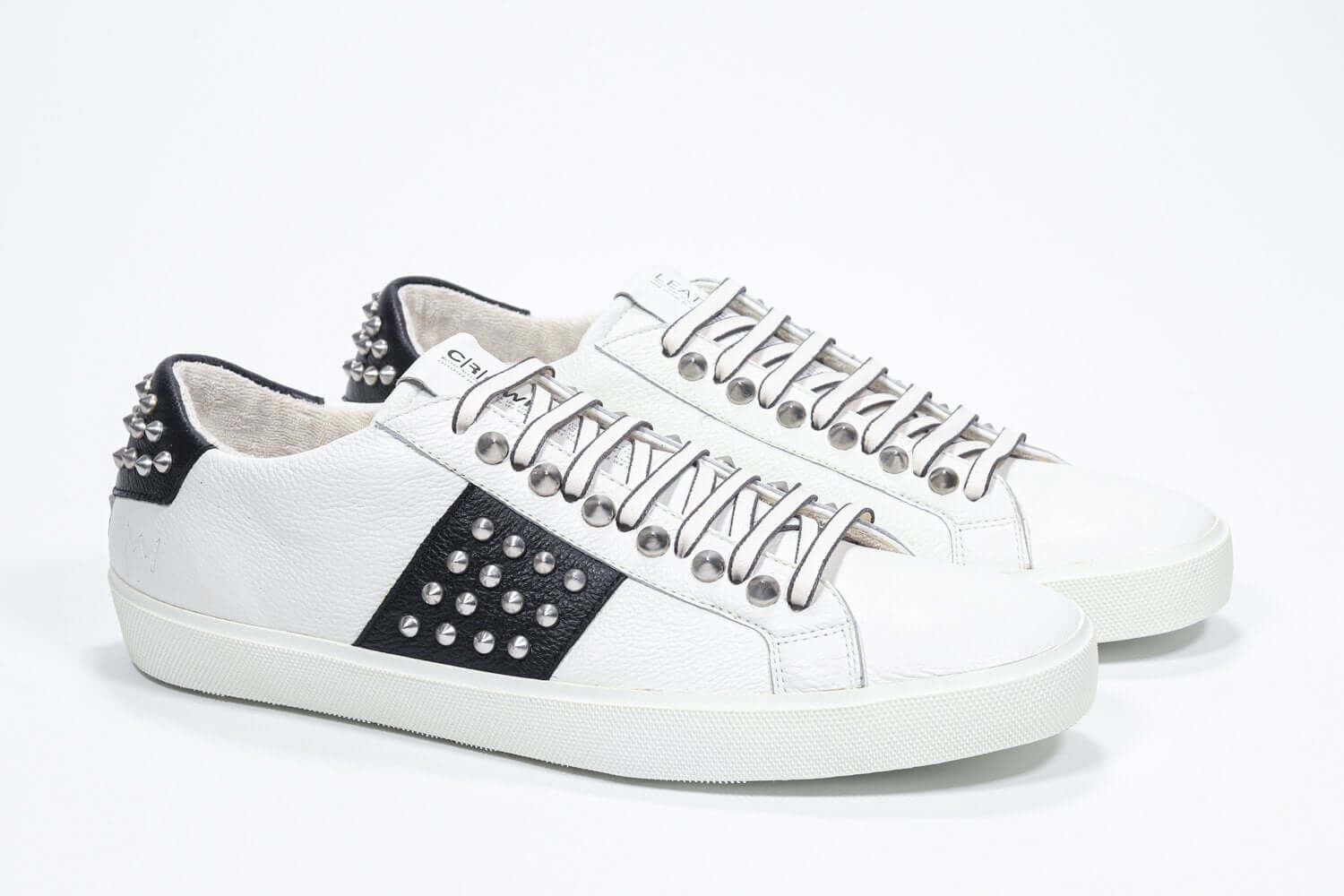 Three quarter front view of low top white and black sneaker. Full grain leather upper with silver studs and white rubber sole.
