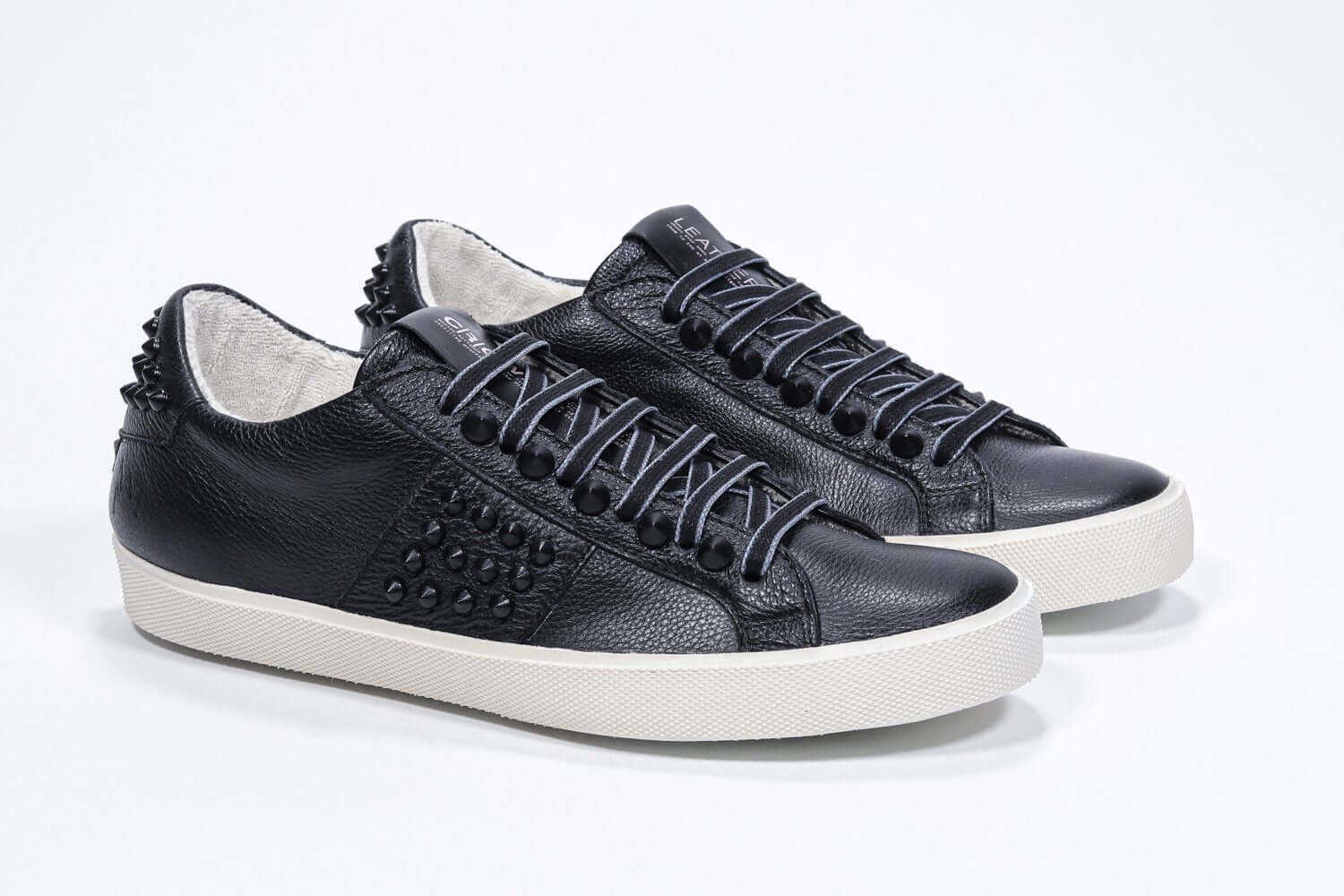 Three quarter view of low top black sneaker. Full grain leather upper with black studs and antique white rubber sole.