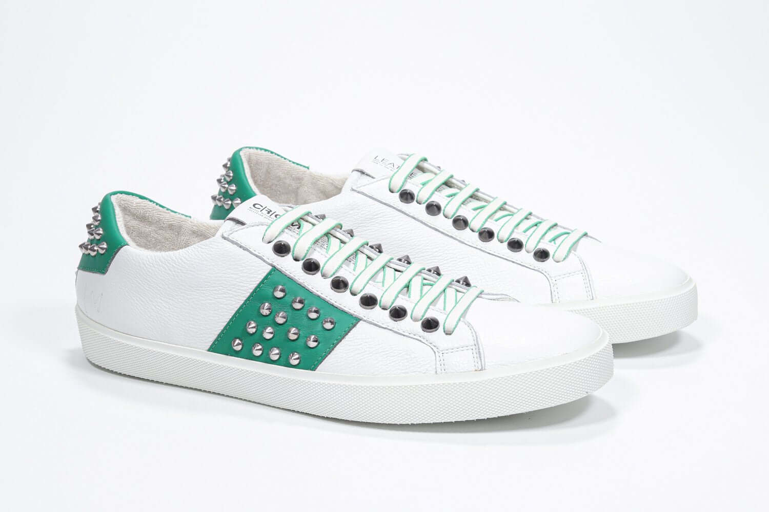 Three quarter front view of low top white and green sneaker. Full grain leather upper with silver studs and white rubber sole.