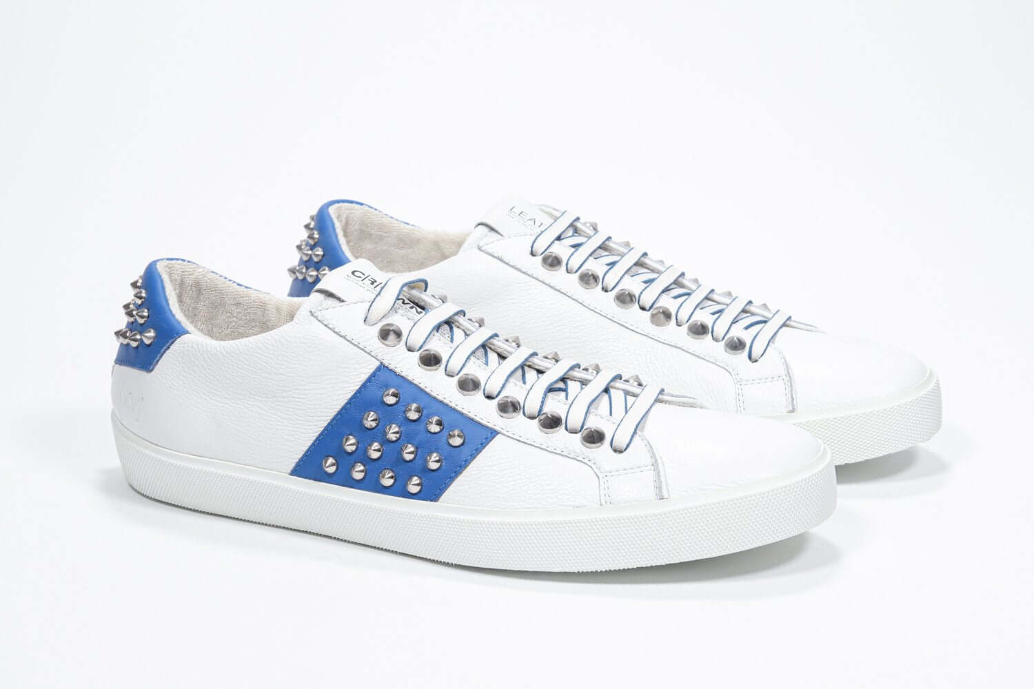 Three quarter front view of low top white and blue sneaker. Full grain leather upper with silver studs and white rubber sole.