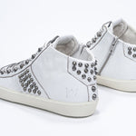 Three quarter back view of mid top white sneaker. Full leather upper with studs, an internal zip and vintage rubber sole.