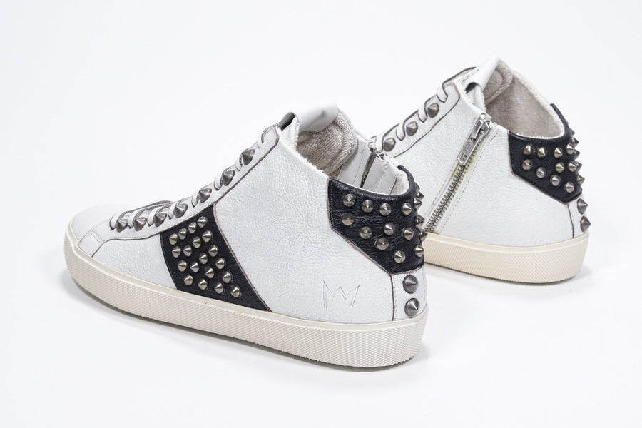 Three quarter back view of mid top white and black sneaker. Full leather upper with studs, an internal zip and vintage rubber sole.