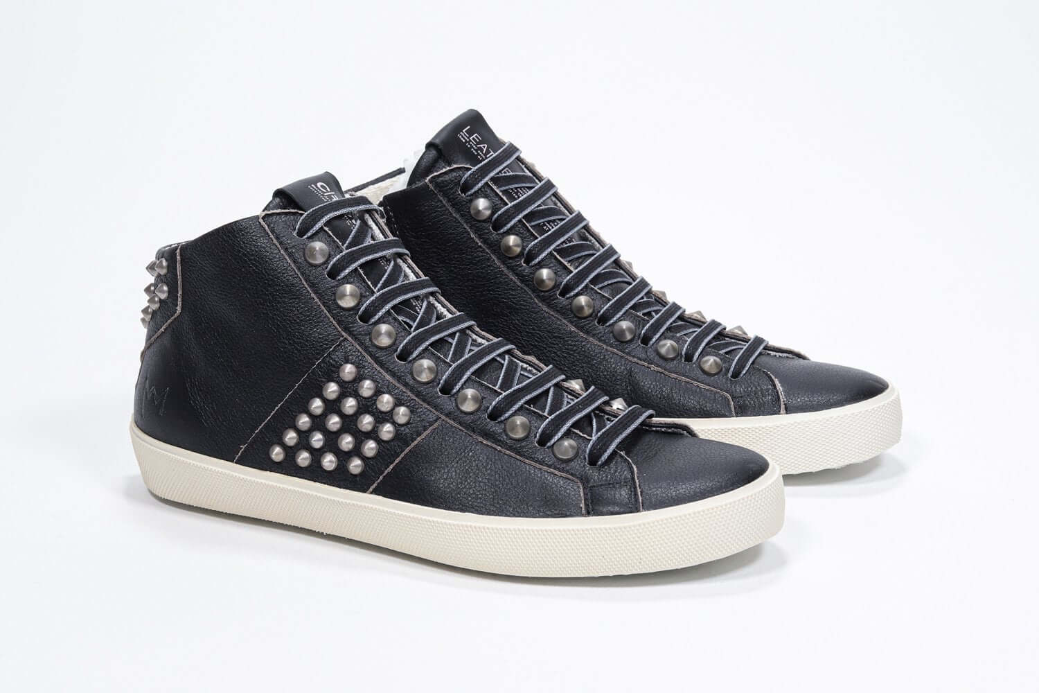 Three quarter front view of mid top black sneaker. Full leather upper with studs, an internal zip and vintage rubber sole.