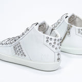 Vue de trois quarts arrière d'une chaussure blanche de taille moyenne sneaker. Tige en cuir avec clous, fermeture à glissière interne et semelle en caoutchouc blanc.