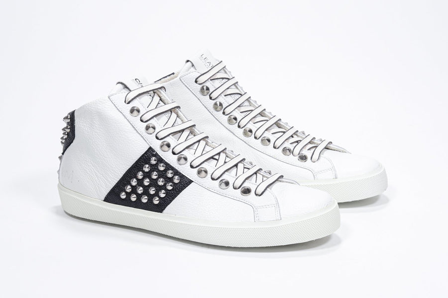 Three quarter front view of mid top white and black sneaker. Full leather upper with studs, an internal zip and white rubber sole.