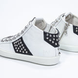 Three quarter back view of mid top white and black sneaker. Full leather upper with studs, an internal zip and white rubber sole.