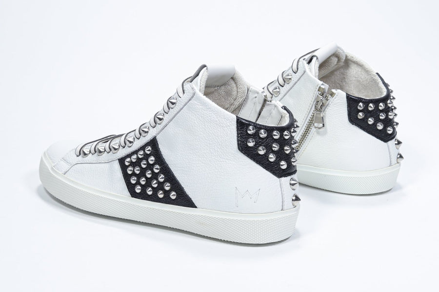 Three quarter back view of mid top white and black sneaker. Full leather upper with studs, an internal zip and white rubber sole.