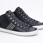 Three quarter front view of mid top black sneaker. Full leather upper with studs, an internal zip and white rubber sole.