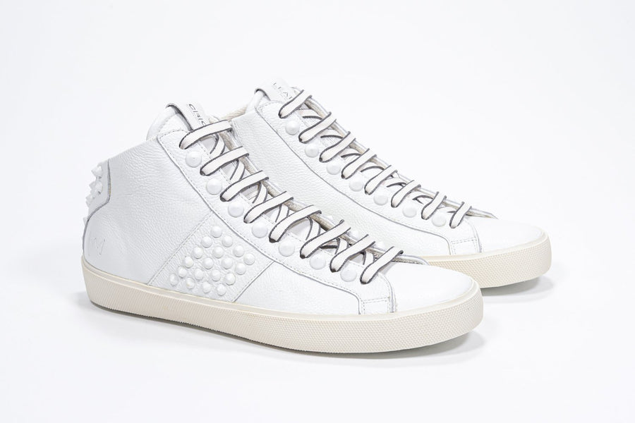Three quarter front view of mid top white sneaker. Full leather upper with studs, an internal zip and vintage rubber sole.