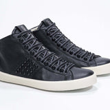 Three quarter front view of mid top black sneaker. Full leather upper with studs, an internal zip and vintage rubber sole.