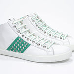 Three quarter front view of mid top white and green sneaker. Full leather upper with studs, an internal zip and white rubber sole.