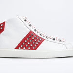 Profilo laterale di mid top bianco e rosso sneaker. Tomaia in pelle con borchie, zip interna e suola in gomma bianca.
