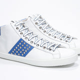 Three quarter front view of mid top white and royal blue sneaker. Full leather upper with studs, an internal zip and white rubber sole.