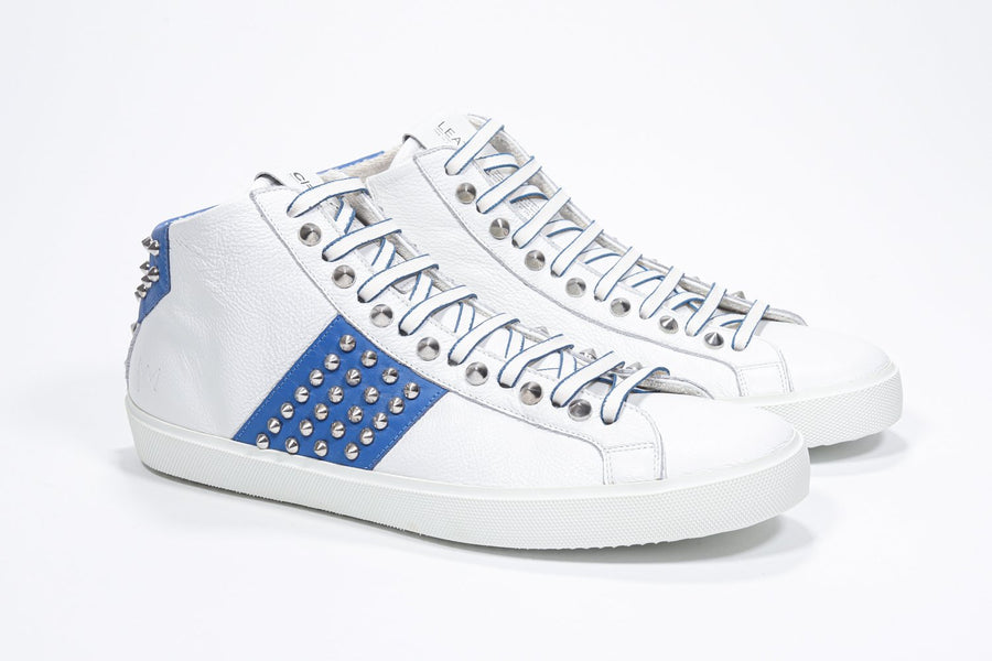 Three quarter front view of mid top white and royal blue sneaker. Full leather upper with studs, an internal zip and white rubber sole.