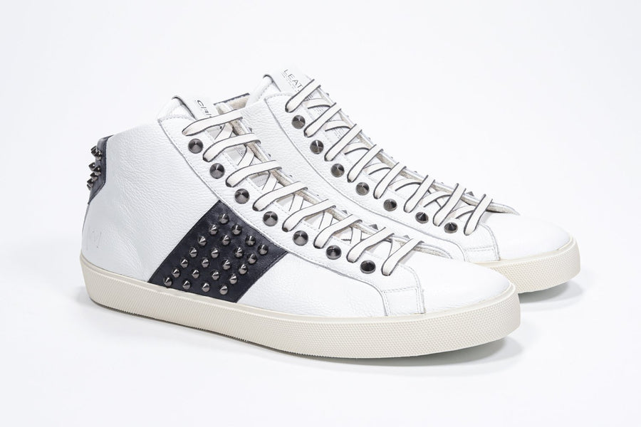 Three quarter front view of mid top white and navy sneaker. Full leather upper with studs, an internal zip and vintage rubber sole.