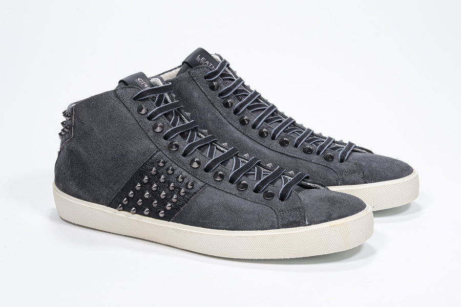 Three quarter front view of mid top dark grey sneaker. Suede and leather upper with studs, an internal zip and vintage rubber sole.