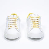 Vue de face de la chaussure blanche sneaker avec des détails jaunes et le logo perforé de la couronne sur la tige. Tige en cuir et semelle en caoutchouc blanc.