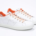 Three quarter front view of low top white sneaker with orange detailing and perforated crown logo on upper. Full leather upper and white rubber sole.