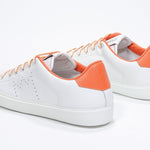 Three quarter back view of low top white sneaker with orange detailing and perforated crown logo on upper. Full leather upper and white rubber sole.
