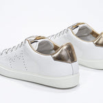 Three quarter back view of low top white sneaker with gold detailing and perforated crown logo on upper. Full leather upper and white rubber sole.
