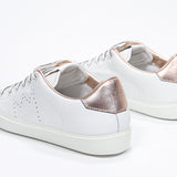 Three quarter back view of low top white sneaker with rose gold metallic detailing and perforated crown logo on upper. Full leather upper and white rubber sole.